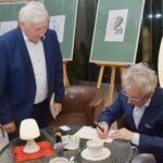 Dwóch mężczyzn w marynarkach rozmawia podczas prezentacji wierszy na sztalugach.