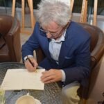 Mężczyzna w garniturze podpisuje dokument leżący na stole, siedząc w fotelu.