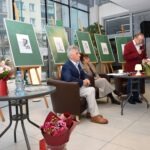 Mężczyźni siedzą w fotelu i przy stole, otoczeni sztalugami z obrazami podczas spotkania Akademii Słowa.