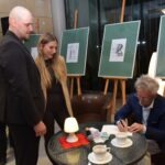 Autor podpisywał książki w towarzystwie ochroniarza i asystentki, w tle widoczne są ekrany z prezentowanymi pracami.