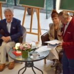 Trzej rozmówcy siedzą przy stoliku z kwiatami i dokumentami podczas spotkania Akademii Słowa.