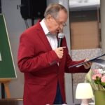 Mężczyzna w czerwonym garniturze przemawia do publiczności, trzymając mikrofon i kartkę z tekstem.