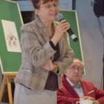 Kobieta w marynarce przemawia do publiczności, trzymając mikrofon przed zieloną tablicą z rysunkiem.