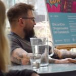 Mężczyzna siedzi przy stole z dzbankiem wody, w tle widoczna tablica reklamowa ChrzanConu.