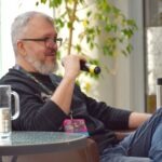 Mężczyzna z brodą pije piwo siedząc przy stoliku na zewnątrz budynku.