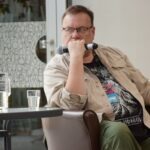 Mężczyzna w okularach siedzi na krześle, trzymając w rękach mikrofon.