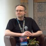 Mężczyzna siedzi na krześle, ubrany w czarny T-shirt i okulary, z identyfikatorem wiszącym na szyi.