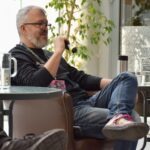 Mężczyzna z brodą siedzi na krześle w sali bibliotecznej, trzymając w ręku aparat fotograficzny.