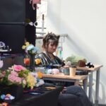 Uczestniczka ChrzanConu w stroju cosplay siedzi przy stoliku otoczonym kwiatami i rekwizytami.