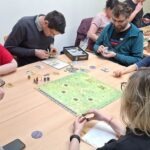 Uczestnicy gry planszowej skupiają się na rozgrywce przy stole pokrytym elementami gry.