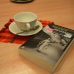 Na drewnianym stole leży książka "Nieczułość" Martyny Bundy obok filiżanki z kawą i łyżeczką na czerwonej serwetce.