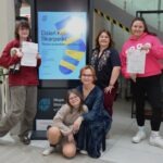 Uśmiechnięta grupa osób prezentuje certyfikaty przed ekranem z informacjami o Dniu Kolorowej Skarpetki.