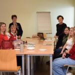 Uczestnicy warsztatów siedzą przy stole w sali konferencyjnej, a część z nich opiera się o niego, obserwując prowadzącą.