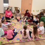 Przedszkolaki siedzą na kolorowej macie podczas zajęć w bibliotece.