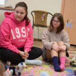 Dwie dziewczynki siedzą na dywanie w pomieszczeniu, jedna z nich nosi okulary korekcyjne i kolorowe skarpetki.