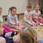 Przedszkolaki siedzą na dywanie podczas zajęć w bibliotece, ubrane w kolorowe skarpetki i wygodne stroje.