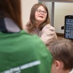 Uśmiechnięta dziewczyna w okularach rozmawia z grupą dzieci podczas warsztatów edukacyjnych.