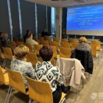 Słuchacze siedzą na żółtych krzesłach w sali konferencyjnej, skierowani w stronę prezentera i ekranu z projekcją.