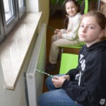Dwie dziewczynki siedzą na krzesłach obok kaloryfera, patrząc w kierunku okna.