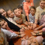 Dzieci uczestniczące w zajęciach bibliotecznych układają dłonie na okrąg wokół stołu.