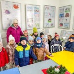 Grupa przedszkolaków stoi przed tablicą z rysunkami, uśmiechając się do kamery w sali bibliotecznej.