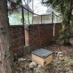 Drewniany domek dla jeża stoi przed ceglanym murem w otoczeniu drzew i liści.