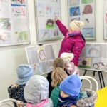 Kobieta wskazuje na portret jeża zawieszony na ścianie, a przed nią stoi grupa dzieci w czapkach.