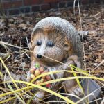 Figurka jeża z kolorowymi koralikami spoczywa wśród suchych liści i gałęzi.