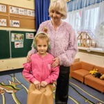 Dziewczynka w różowej bluzie i czapce z jeżem stoi obok starszej kobiety na tle tablicy z pracami dziecięcymi.