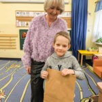 Chłopiec trzyma brązowy papierowy worek, a za nim stoi uśmiechnięta starsza kobieta w fioletowym swetrze.