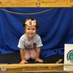 Dziecko z opaską w kształcie jeża uśmiecha się do kamery podczas zajęć edukacyjnych.