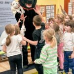 Dzieci z przedszkola podnoszą ręce do małego jeża trzymanego przez osobę przebraną w kostium czarnego psa.