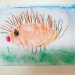 Narysowane kredkami jeżyk ma pomarańczowe ciało pokryte kolcami, różową noskę i zieloną trawę na podkładzie.