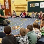 Grupa przedszkolaków słucha opowieści osoby prowadzącej warsztaty ekologiczne o jeżu, siedząc na dywanie z motywem leśnym.