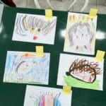 Na zielonym stole przymocowane żółtymi taśmami znajdują się rysunki portretów jeży wykonane przez dzieci.
