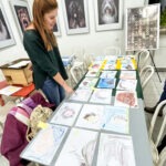 Na stole ułożone są prace plastyczne dzieci przedstawiające jeże, a w tle widoczne są ramki z innymi ilustracjami.