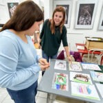 Dwie kobiety przeglądają prace konkursowe przedstawiające jeże, rozłożone na stole w bibliotece.