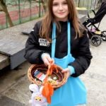 Dziewczynka trzyma wiklinowy koszyk wypełniony kolorowymi jajkami oraz pluszową zabawką.