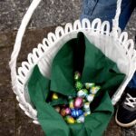 W wiklinowym koszyku wyłożonym zieloną tkaniną leżą kolorowe jajka czekoladowe.