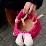 Dziecko trzyma ręką różowy, robiony na drutach koszyczek z białymi pomponami.