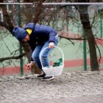 Chłopiec w niebieskiej kurtce pochyla się, zbierając jajko do białego koszyka na placu zabaw.