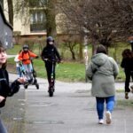 Na ścieżce parkowej grupa osób przemieszcza się różnymi środkami transportu, w tym z wózkiem dziecięcym i hulajnogą elektryczną.