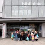 Grupa dzieci z rodzicami stoi przed budynkiem biblioteki, trzymając koszyczki.