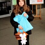 Dziewczynka trzyma koszyk z pluszakiem oraz niebieską torbę, prawdopodobnie zebranymi słodyczami.