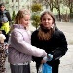 Dwie dziewczynki w kurtkach podają sobie koszyczek z jajkami podczas polowania wielkanocnego.