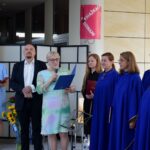 Grupa osób stoi przed budynkiem biblioteki podczas uroczystości, w tym kobiety w niebieskich szatach i osoba mówiąca do mikrofonu.