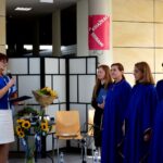 Kobieta przemawia podczas uroczystości w obecności chóru w niebieskich szatach, z bukietami słoneczników i flagą na tle.