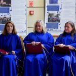 Trzy kobiety w niebieskich szatach siedzą na krzesłach i trzymają nuty.