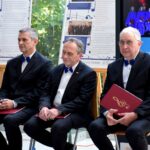 Trzech mężczyzn w garniturach siedzi na krzesłach podczas uroczystości, za nimi widoczna dekoracja i chór.