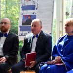 Trzej mężczyźni siedzą na krzesłach podczas wydarzenia, trzymając czerwone teczki, a za nimi widać plakaty i okna z widokiem na zielony teren.